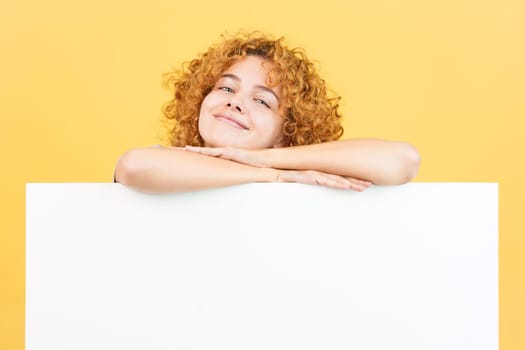 Studio image with yellow background of a relaxed woman with curly hair leaning on a blank panel