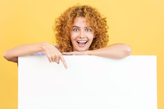 Studio image with yellow background of a smiley woman with curly hair pointing to a blank panel