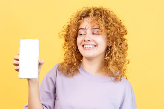Studio image with yellow background of a young and beauty smiley woman showing the blank screen of the mobile