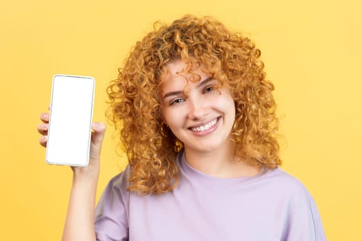Studio image with yellow background of a smiley woman looking at camera while holding a mobile with the blank screen