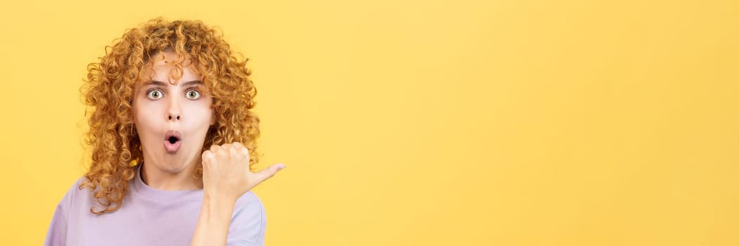 Panoramic studio image with yellow background of a young woman with curly hair pointing surprised to the side