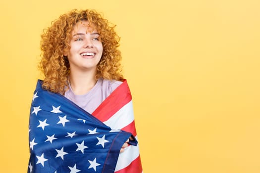 Studio image with yellow background of a distracted and dreamy woman with curly hair wrapping with a north america national flag
