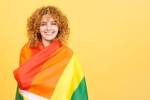 Studio image with yellow background of a woman wrapped with a lgbt flag looking at camera
