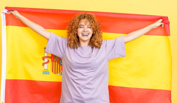 Studio image with yellow background of a happy beauty woman with curly hair raising a spanish national flag