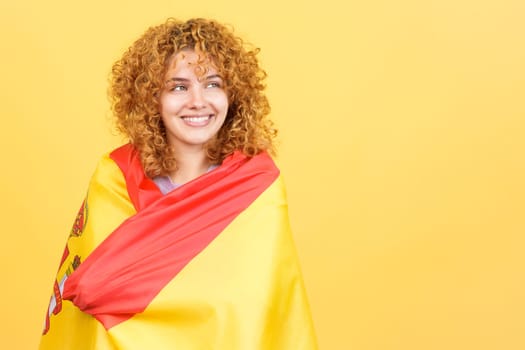 Studio image with yellow background of a happy woman looking aside distracted while wrapping with a spanish national flag