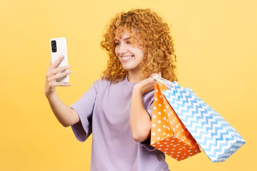 Studio image with yellow background of a happy young woman with curly hair taking a selfie while holing shopping bags