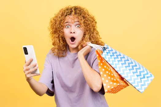 Studio image with yellow background of a surprised woman using the mobile while holing shopping bags