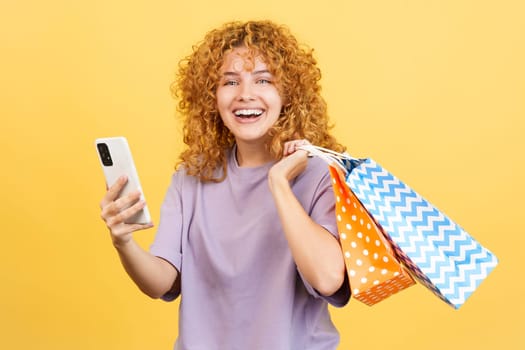 Studio image with yellow background of a young smiley woman with curly hair using the mobile while holing shopping bags