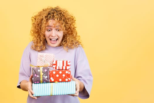 Studio image with yellow background of an excited young woman with curly hair and casual clothes receiving many gifts