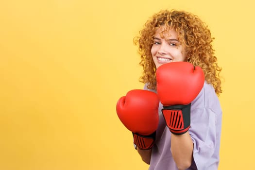 Studio image with yellow background of a cool woman with curly hair using boxing gloves while smiling at the camera