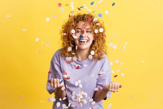 Studio image with yellow background of a young and cheerful woman with curly hair surrounded by confetti in the air