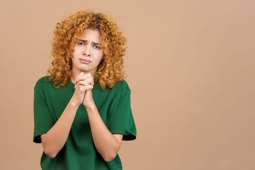 Studio photo of woman beggong with curly hair and a worried face, praying with her hands clasped while looking into the camera