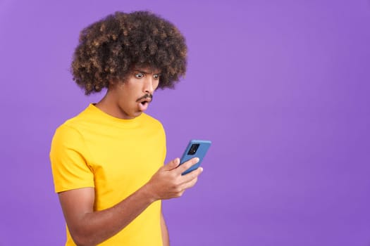 Surprised african man with curly hair looking the screen of a mobile in studio