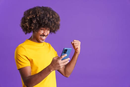 Excited african man with curly hair celebrating while using the mobile phone in studio