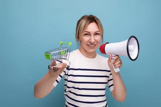 blond attractive young woman in casual attire speaks using a megaphone while holding a supermarket trolley on a blue background.