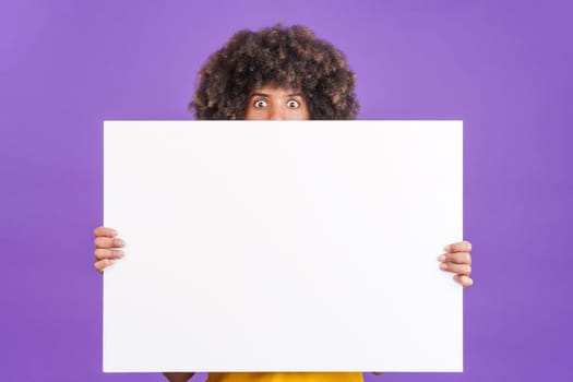 Surprised african man with curly hair hiding behind a blank panel in studio