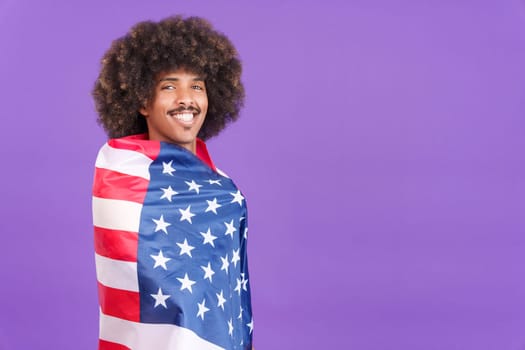 Happy african man with curly hair wrapping with a north america national flag while looking at camera in studio