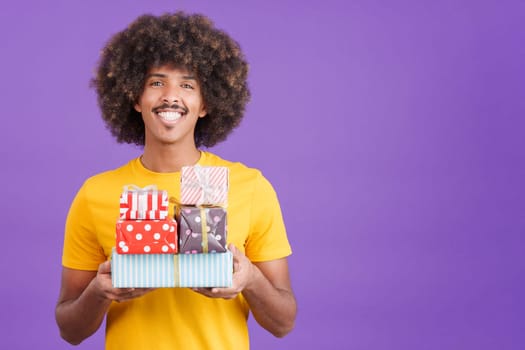 Smiley african man with curly hair holding many gifts while looking at the camera in studio
