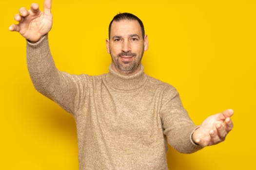 Bearded Hispanic man wearing a beige turtleneck in a friendly attitude looking at the camera smiling with open arms for a hug. Cheerful expression embracing happiness. Isolated on yellow background