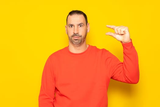 Handsome hispanic man with beard a red sweater gesturing with his hand making a small size sign with fingers looking and camera. Measurement concept. Isolated on yellow background.