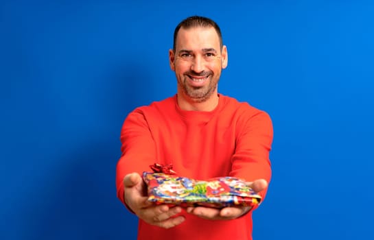Bearded Hispanic man in his 40s wearing a red sweater delivering a birthday present to his beloved, he smiles with satisfaction as his partner has been pleasantly surprised. Isolated blue background