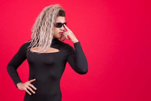 Transgender person dressed as drag queen wearing sunglasses posing with rebel attitude in studio with red background
