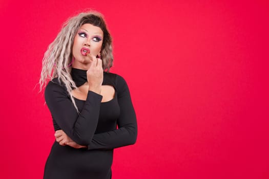 Distracted transgender person dressed as drag queen applying lipstick in studio with red background