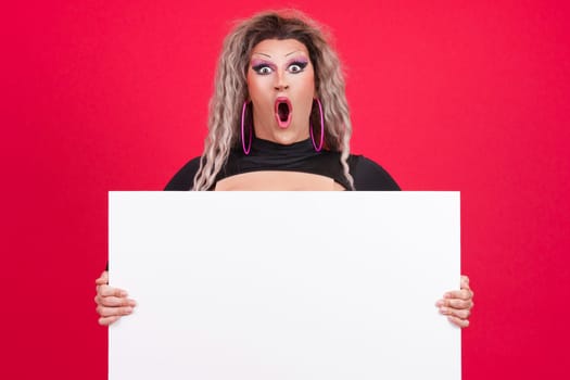 Surprised transgender person with make up opening the mouth while holding a blank panel in studio with red background