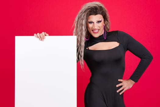 Smiley transgender person dressed as drag queen and with make up holding a blank panel in studio with a red background