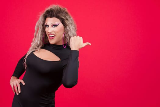 Transgender person dressed as drag queen smiling at camera and pointing to the side in studio with red background