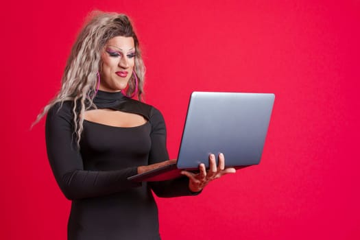 Happy transgender person dressed as drag queen standing and using a laptop in studio in red background