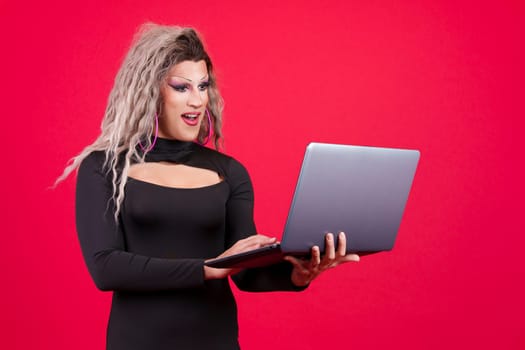 Surprised transgender person dressed as drag queen standing and using a laptop in studio in red background