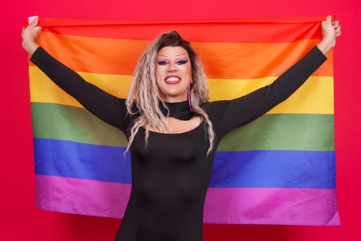 Happy transgender person dressed as drag queen raising a lgbt rainbow flag in studio with red background
