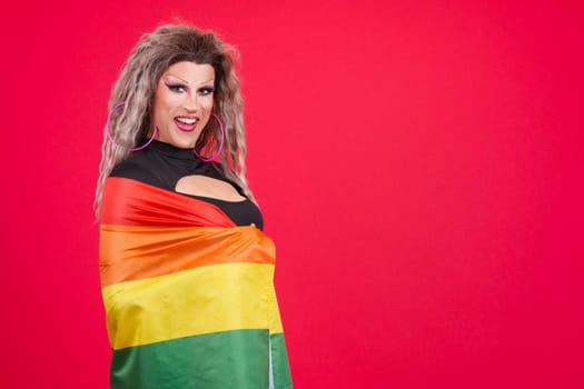 Happy transgender person dressed as drag queen looking at camera while wrapping with a rainbow lgbt flag in studio with red background