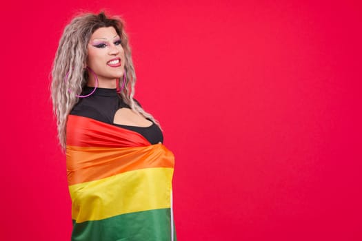Proud transgender person dressed as drag queen wrapping with a rainbow lgbt flag in studio with red background