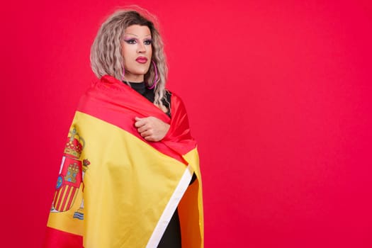 Serious transgender person dressed as drag queen wrapping with a spanish flag in studio with red background
