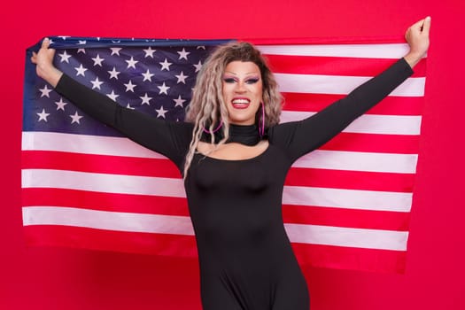 Happy transgender person dressed as drag queen raising a north america national flag in studio with a red background
