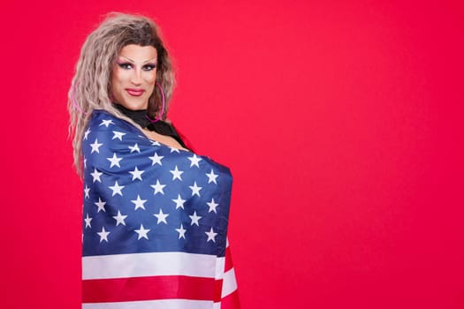 Happy transgender person dressed as drag queen smiling at camera while wrapping with a north america national flag in a red background