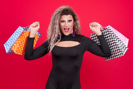 Sensual transgender person dressed as drag queen smiling at the camera while showing shopping bags in studio with red background