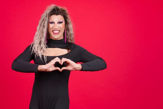 Drag queen man in black dress and make up representing a heart in the shape of fingers in studio with red background