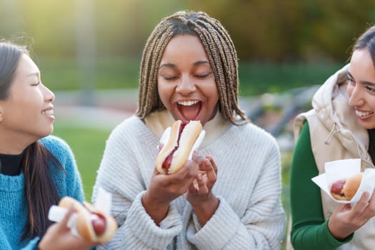 Three multiethnic friends eating hot dogs in a park