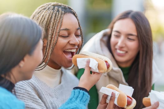 Three multicultural smiling friends eating hot dog in a park