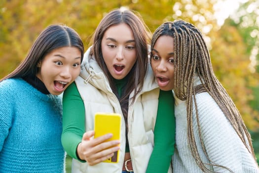 Three surprised multicultural friends using a mobile in a park