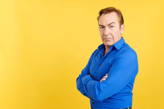 Serious caucasian mature man turning to face the camera while standing with arms crossed in studio with yellow background