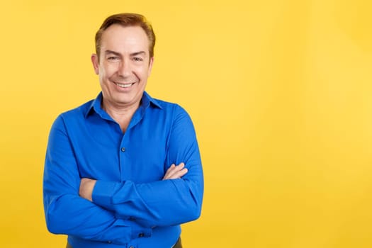 Mature caucasian man smiling at the camera with arms crossed in studio with yellow background