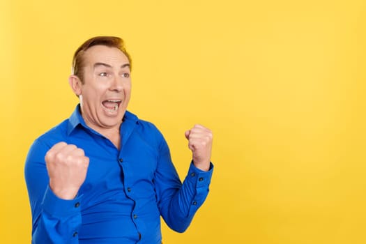 Happy mature man celebrating raising the fists in studio with yellow background