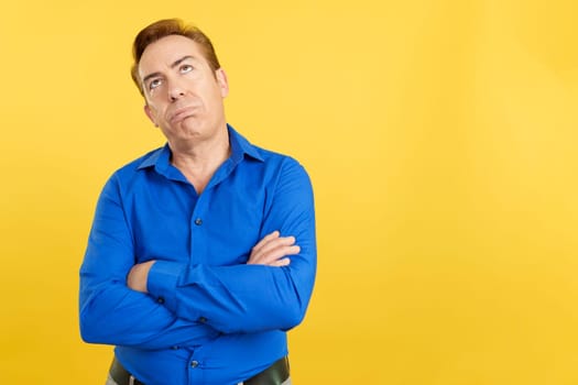 Bored mature man looking up with arms crossed in studio with yellow background