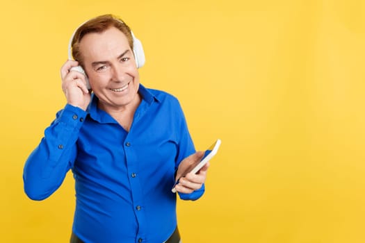 Mature man looking at camera while listening to music with headphones and mobile in studio with yellow background