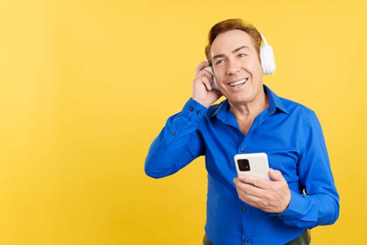 Smiley mature man listening to music with the mobile and headphones in studio with yellow background