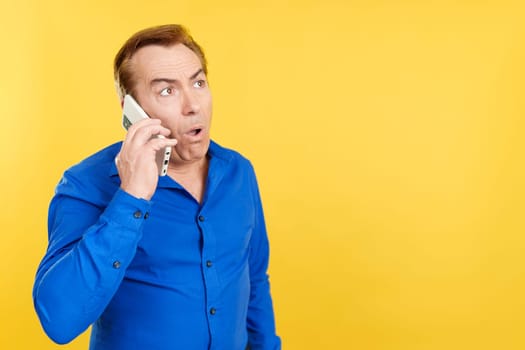 Surprised mature man talking to the mobile in studio with yellow background